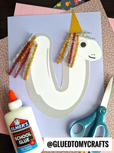 A pipe cleaner unicorn craft made from paper, with a gold horn, googly eye, and colorful mane on a purple background. Elmers glue, scissors, and patterned paper are nearby. Perfect for “U is for Unicorn” projects! @GLUEDTOMYCRAFTS is shown at the bottom.