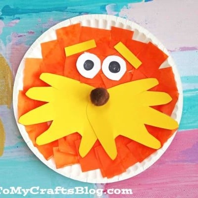 A paper plate Lorax craft features an orange tissue paper face, big yellow mustache and eyebrows, googly eyes, and a brown pom-pom nose on a colorful painted background.