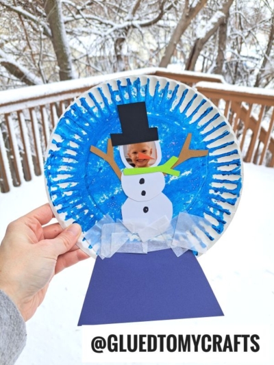 A hand holds a paper plate snowman snow globe craft with a child’s photo as the snowman’s face, blue painted background, stick arms, black hat, green scarf, and tissue paper snow. A snowy outdoor scene is visible in the background.