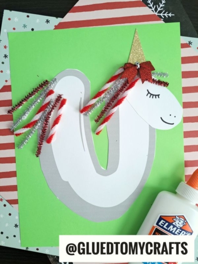 A pipe cleaner unicorn craft made from paper and shiny pipe cleaners sits on a green background. The unicorn has a gold horn, red bow, closed eyes, and Elmer’s glue is nearby—perfect for a U is for Unicorn project!.
