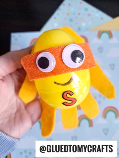 A hand holds a yellow plastic egg superhero decorated with an orange felt mask and cape, googly eyes, and an S on its front. The background features pastel rainbows and clouds. Text reads @GLUEDTOMYCRAFTS.