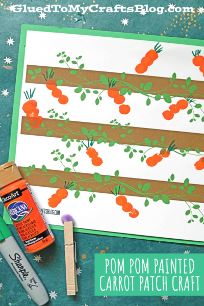 A children’s craft project featuring a pom pom painted carrot patch made with orange pom-pom carrots and green leafy tops on brown paper strips, with a paint bottle, Sharpie marker, and clothespin on the side.