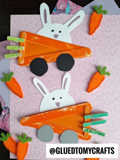 Two bunny crafts made from orange-painted popsicle sticks shaped like popsicle stick carrot cars, with bunny faces peeking over the top and green clothespins as carrot tops. Decorative carrots and @GLUETOMYCRAFTS logo are also visible.