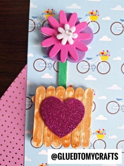 A handmade craft featuring a pink and white foam flower on a green stem, set in a popsicle stick flower pot adorned with a glittery pink heart. The background showcases bicycle patterns and pink polka dot paper.