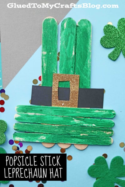 A popsicle stick leprechaun hat with a gold glitter buckle and black band sits amid confetti and glittery shamrocks, all set against a vibrant blue background.