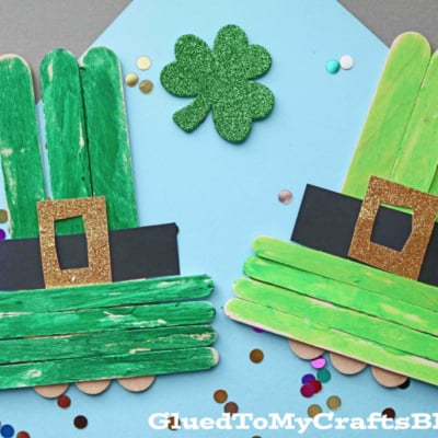 Two popsicle stick leprechaun hats, crafted from green-painted sticks and adorned with gold glitter buckles and black bands, are placed on a blue background alongside a green glitter shamrock and colorful confetti.