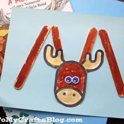A children’s book, If You Give a Moose a Muffin, is shown with a crafty popsicle stick moose made from googly eyes, paper antlers, and a paper face on a sparkly surface.