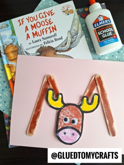 Children’s craft supplies and books displayed: a book titled “If You Give a Moose a Muffin,” a bottle of Elmer’s School Glue, and a popsicle stick moose craft made from paper and sticks on pink paper.