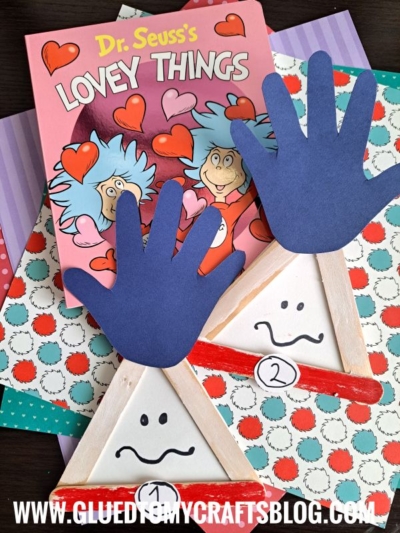 Colorful craft scene featuring Dr. Seuss’s Lovey Things book, patterned paper, blue handprint cutouts, handmade triangle crafts with faces and numbers, and a Popsicle Stick Thing 1 & 2 project. Website gluedtomycraftsblog.com appears at the bottom.