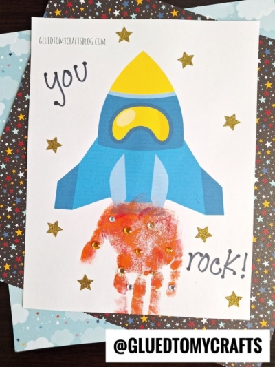 A blue rocket ship soars above a red handprint that looks like rocket flames, creating a charming Handprint Rocket Keepsake. Gold glitter stars and the words "you rock!" decorate the scene on a star-patterned background.