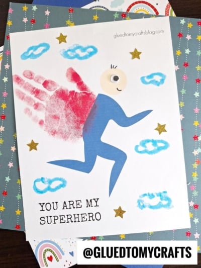 A handmade card featuring a blue handprint superhero figure with a red cape, cartoon face, clouds, gold stars, and the text YOU ARE MY SUPERHERO. The @GLUEDTOMYCRAFTS watermark is at the bottom.