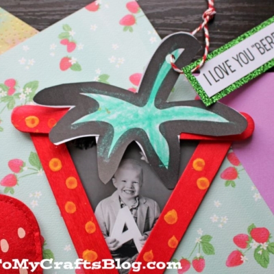 A popsicle stick strawberry photo frame, painted and shaped like a berry with a green top and a tag reading I LOVE YOU BERRY MUCH, is displayed alongside paper strawberries on a cheerful polka dot background.