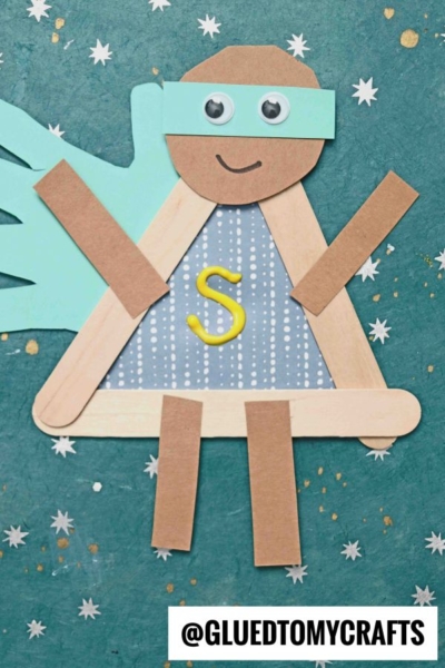 Create a popsicle stick superhero with brown paper, a blue mask and cape, googly eyes, and a yellow “S” on the chest, displayed against a starry teal background.