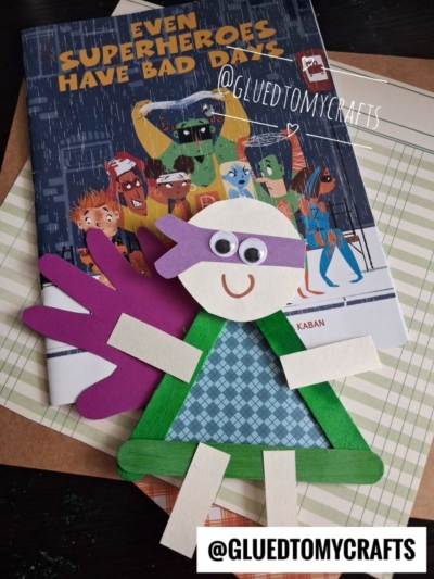 A handmade popsicle stick superhero craft with googly eyes and a purple mask lies on a book titled Even Superheroes Have Bad Days, featuring illustrated superheroes on the cover.