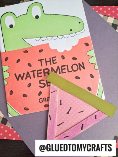 A children’s book titled The Watermelon Seed with a cartoon crocodile and watermelon cover is displayed next to a handmade Popsicle Stick Watermelon Slices bookmark, on a colorful background. @GLUEDTOMYCRAFTS is printed at the bottom.