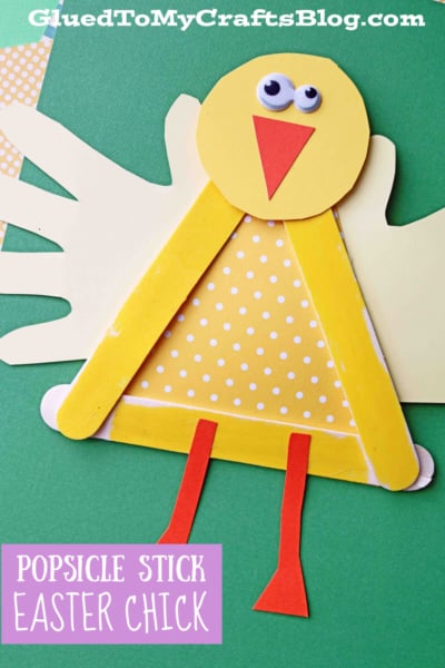 A popsicle stick baby chick craft made from yellow sticks, paper wings, a polka dot triangle body, orange beak and legs, and googly eyes is displayed on a green background.