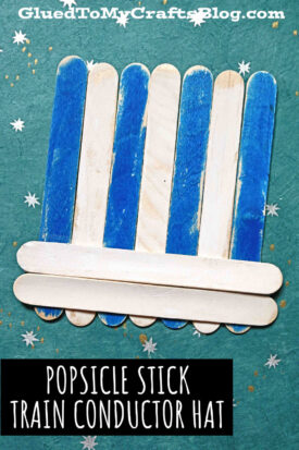 A popsicle stick train conductor hat, crafted from blue and plain sticks, sits on a green background sprinkled with tiny white stars. Perfect as a Christmas conductor hat craft. Text reads, Popsicle Stick Train Conductor Hat.