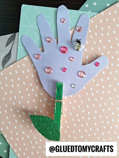A paper handprint flower decorated with pink and clear sequins is attached to a green clothespin stem with a glittery leaf, placed on pastel paper. The bottom has @GLUEDTOMYCRAFTS text.