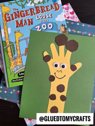 A handmade paper handprint giraffe card with brown spots and googly eyes rests on green paper, next to the book The Gingerbread Man Loose at the Zoo. The @GLUEDTOMYCRAFTS handle is visible.