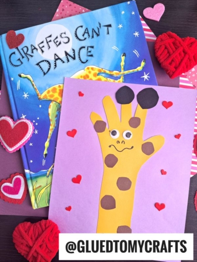 A children’s book titled “Giraffes Can’t Dance” sits next to a colorful paper handprint giraffe craft, made from a yellow handprint on purple paper, decorated with brown spots, googly eyes, and surrounded by red hearts.