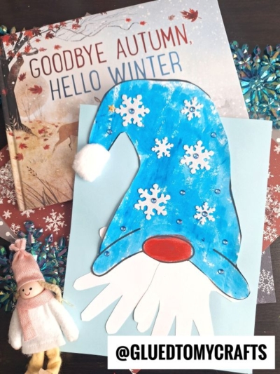 A handmade paper handprint gnome with a handprint beard, blue paper hat, and snowflake decorations sits on a book titled Goodbye Autumn, Hello Winter. Nearby are a small winter doll and blue snowflake ornaments.
