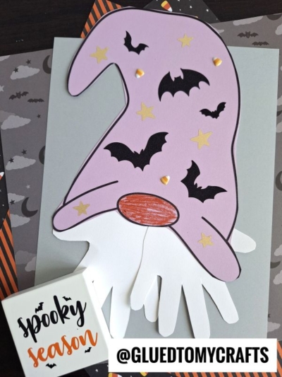 A purple paper witch hat with bat and star cutouts is glued over two white paper hands, creating a whimsical paper handprint gnome with a handprint beard on gray paper. "Spooky season" and @GLUEDTOMYCRAFTS are printed on cards in the image.