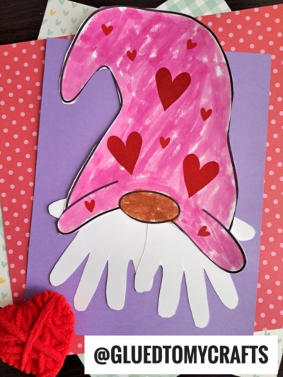 A Valentines Day paper handprint gnome craft featuring a pink hat with red hearts, a brown nose, and a handprint beard made from cut-out shapes on purple paper, surrounded by fun patterned backgrounds.
