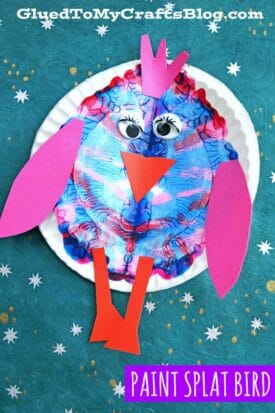 A colorful paper plate paint splat bird craft features a blue and pink paint-splattered body, pink paper wings, feet, and crown, googly eyes, and an orange beak on a teal background with white stars. Text reads “PAINT SPLAT BIRD.”.