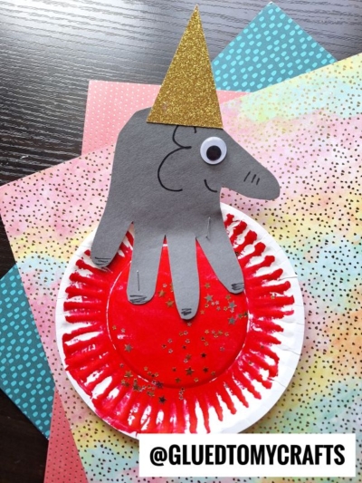 A circus elephant craft for kids made from a gray handprint cutout with a googly eye and gold party hat, sitting on a red paper plate decorated with gold stars, and set on colorful patterned paper.