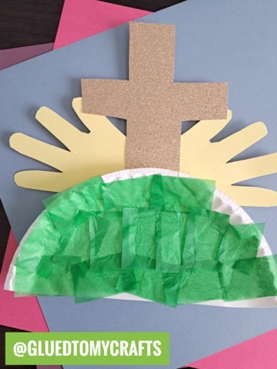 A children's craft features a gold glitter cross, yellow paper rays, and a green tissue paper hill on a blue background—perfect for a paper plate Easter scene. The Instagram handle @gluedtomycrafts appears at the bottom.