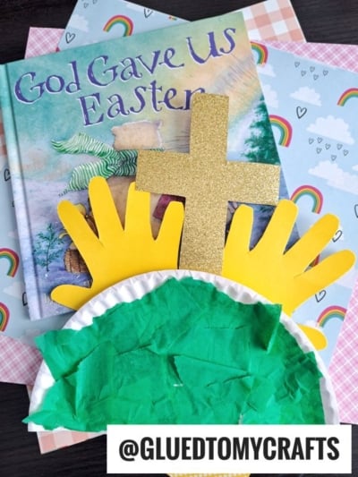 A children's book titled God Gave Us Easter sits on rainbow-patterned paper next to a paper plate Easter scene made of yellow handprints, a glittery gold cross, and green tissue paper. @GLUEDTOMYCRAFTS is at the bottom.