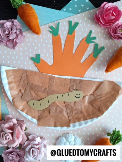 A kids’ craft featuring a paper plate handprint carrot, a smiling paper worm, fake carrots, and pink flowers on a polka dot background. @GLUEDTOMYCRAFTS is written at the bottom.