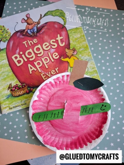 A children’s book titled The Biggest Apple Ever lies next to a paper plate apple w/worm craft painted red to resemble an apple, with green and brown construction paper pieces, a smiley face, and a playful popsicle stick worm peeking out. @GLUEDTOMYCRAFTS is printed twice.