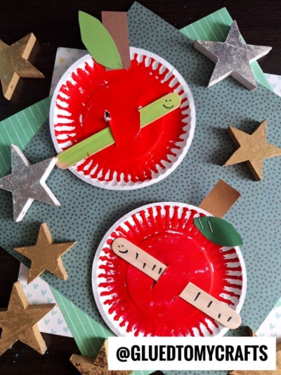 Two paper plate apple w/worm crafts painted red, each with a popsicle stick worm—one green and smiling, the other tan with black dots. Gold and silver stars surround the apples. @GLUEDTOMYCRAFTS text sits at the bottom.
