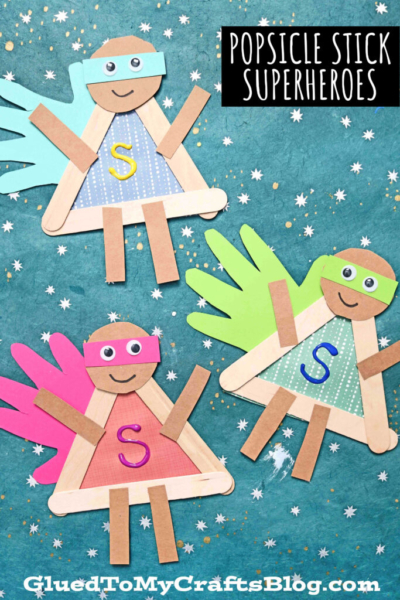 Three colorful popsicle stick superhero crafts made from paper and googly eyes are displayed on a blue starry background. Each figure has a mask, cape, and a large letter S on its chest.
