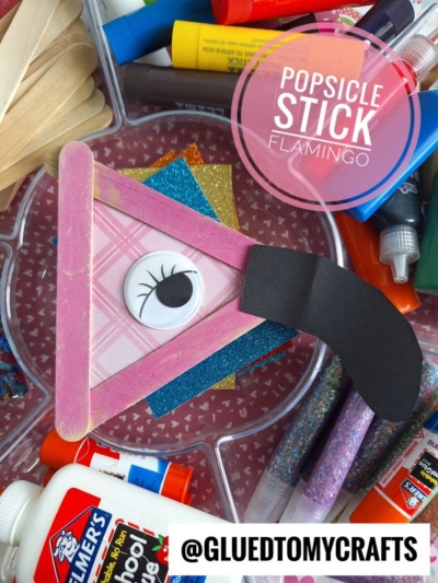 A handmade craft stick flamingo features a popsicle stick flamingo head with a large googly eye, black paper beak, and pink plaid paper, surrounded by craft supplies like glue, markers, glitter, and colored paper. Text: “Popsicle Stick Flamingo.”.