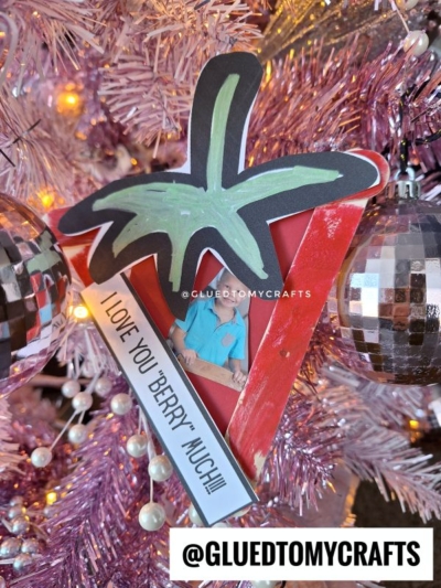 A handmade popsicle stick strawberry ornament featuring a child’s photo and the message “I LOVE YOU ‘BERRY’ MUCH!!!” hangs on a pink Christmas tree with metallic ball ornaments. “@GLUEDTOMYCRAFTS” appears at the bottom.