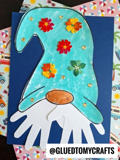 A colorful paper handprint gnome with a blue hat, red, yellow, and green flower cutouts, a white handprint beard, and a brown nose is displayed on a navy blue sheet with patterned paper beneath.