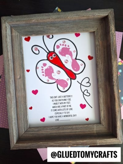A framed artwork of a footprint butterfly made from pink and red painted handprints, decorated with small red hearts. Beneath the butterfly is a heartfelt poem about love and handmade art. The handle @GLUEDTOMYCRAFTS is at the bottom.