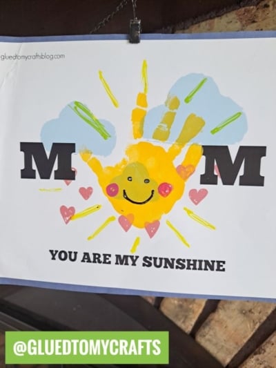 A handmade handprint mom card spells MOM with a yellow handprint as the O, featuring a smiley face, pink hearts, and blue skies. Below, it reads MOM YOU ARE MY SUNSHINE. A green label says @GLUEDTOMYCRAFTS.