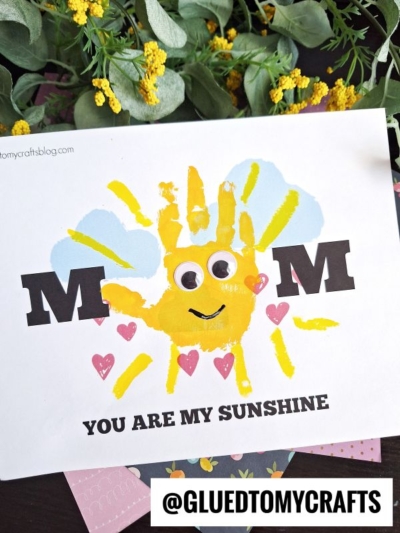 A handmade handprint mom card with the word MOM and a yellow handprint forming a sun, with a smiling face and pink hearts. The text below reads, MOM YOU ARE MY SUNSHINE. Yellow flowers decorate the background.
