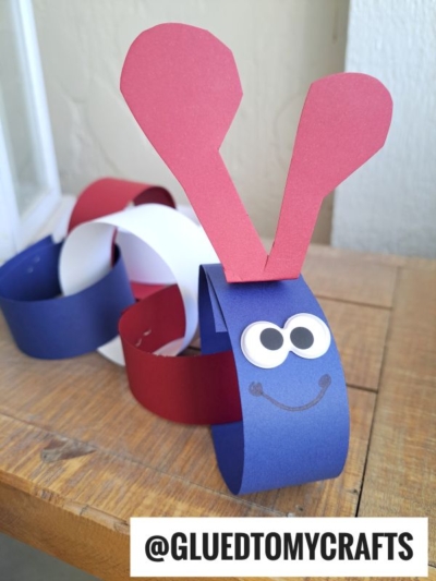 A paper chain caterpillar made of red, white, and blue rings with a blue head, googly eyes, a smiling mouth, and red antennae. Text @GLUEDTOMYCRAFTS is at the bottom. Perfect for your next paper caterpillar craft!.
