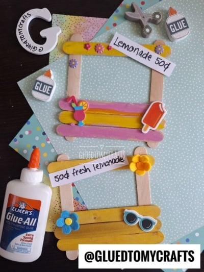 Craft supplies surround two popsicle stick lemonade stands on pastel polka dot papers, decorated with paper flowers, sunglasses, and lemonade signs. Nearby are a bottle of Elmer’s glue and fun craft embellishments.