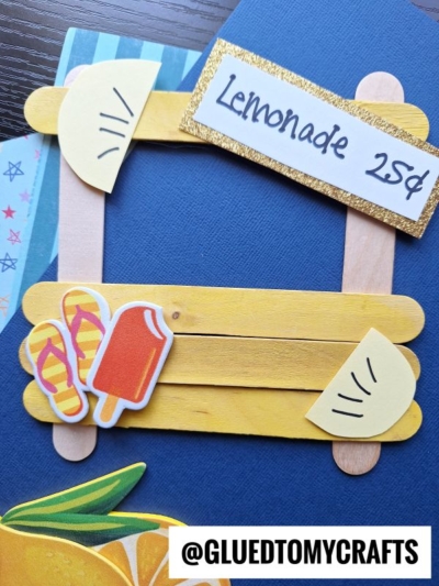 A popsicle stick lemonade stand craft decorated with paper lemons, flip-flops, an ice pop, and a “Lemonade 25¢” sign on a blue background. @GLUEDTOMYCRAFTS appears at the bottom.