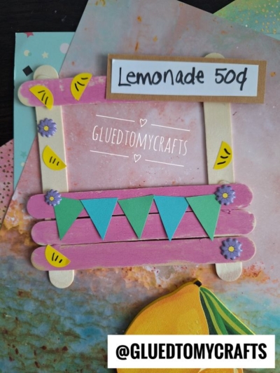 A handmade popsicle stick lemonade stand craft with pink and yellow sticks, blue pennant banners, and lemon slice decorations. A sign reads Lemonade 50¢. Colorful papers in the background. @GLUEDTOMYCRAFTS at the bottom.