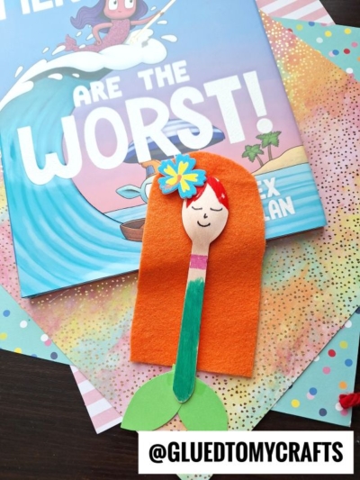 A handmade wooden spoon mermaid craft idea for kids, featuring orange felt hair, a green paper tail, and a drawn face—displayed on a colorful book and patterned paper background with the text @GLUEDTOMYCRAFTS.