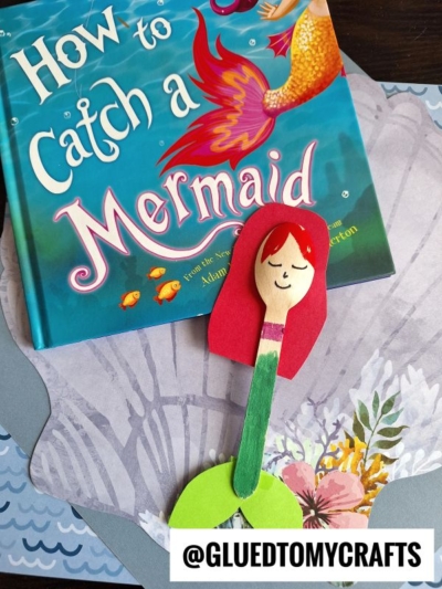 A children's book titled How to Catch a Mermaid sits next to a wooden mermaid spoon craft, featuring red paper hair, a drawn-on face, and a green paper tail. The handle has a pink sticker—a fun mermaid craft idea for kids.