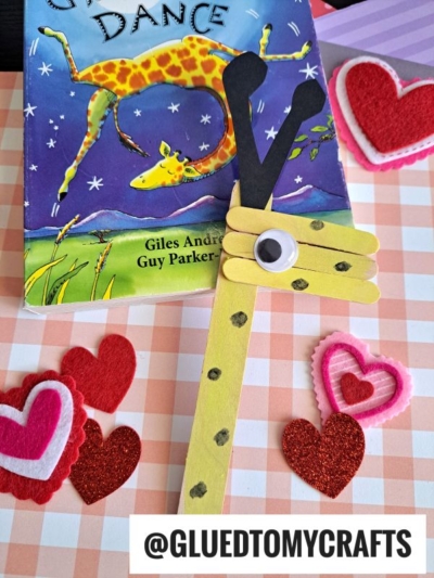 A popsicle stick giraffe craft with a googly eye sits on a pastel checkered surface in front of the book Giraffes Can't Dance. Felt and glitter heart shapes decorate the scene. Text reads @GLUEDTOMYCRAFTS.