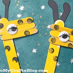 Two popsicle stick giraffe crafts, painted yellow with black paper ears, black spots, and googly eyes, stand against a teal background dotted with white star designs. Their playful, cartoon-like faces add extra charm.