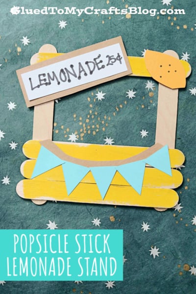 A handmade popsicle stick lemonade stand crafted from sticks, paper, and cardboard features a LEMONADE 25¢ sign and a blue pennant banner, displayed on a green background with white star decorations.